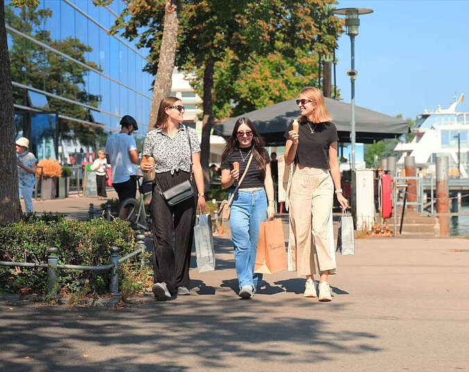 Screenshot aus Film: Drei junge Frauen beim Shoppen mit Tüten. Sie schlendern entlang der Uferpromenade.