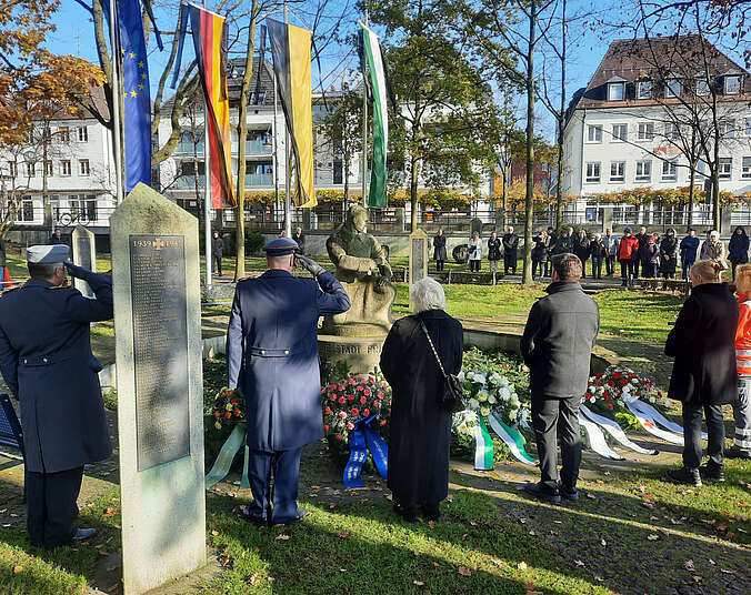 Menschen stehen um Kriegerdenkmal an der Uferpromenade; es liegen Kränze vor dem Denkmal