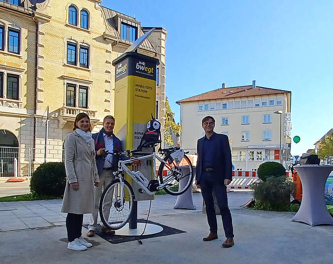 Drei Personen stehen vor gelber Säule an der ein Fahrrad hängt.